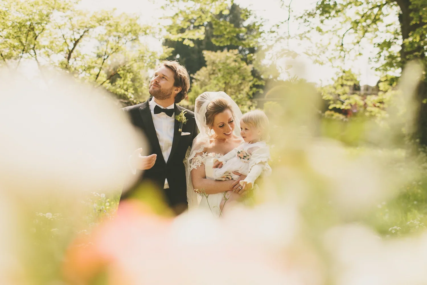 A wedding scene outdoors with a bride, groom, and a young girl surrounded by trees and flowers.