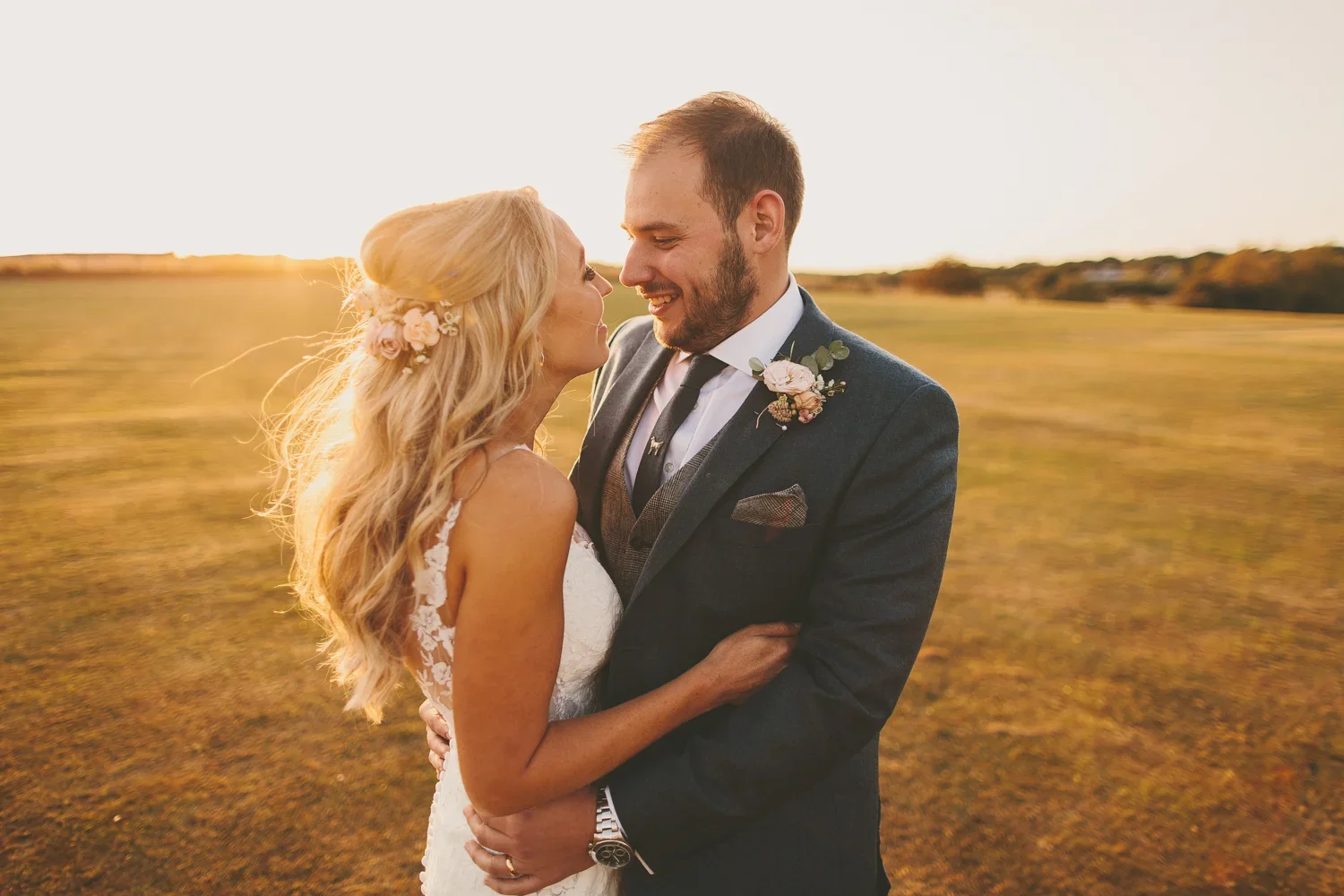 A bride and groom sharing a romantic moment outdoors during sunset, embracing and smiling at each other in a field.