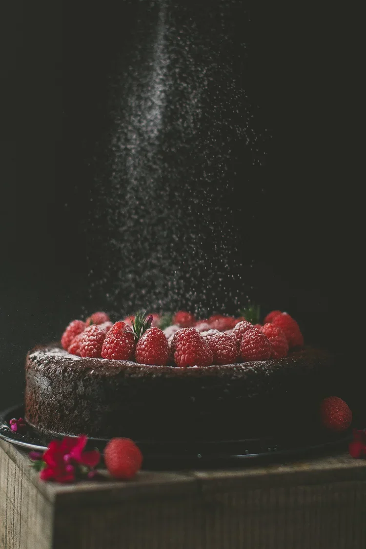 Chocolate cake decorated with fresh raspberries, with powdered sugar sprinkled from above, set on a dark background with some flowers nearby.