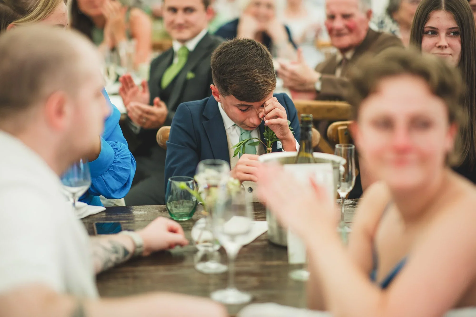 A young boy in a blue suit is sitting at a dining table, pinching his nose and holding a small branch in front of him, with a group of smiling people clapping and looking on in the background.