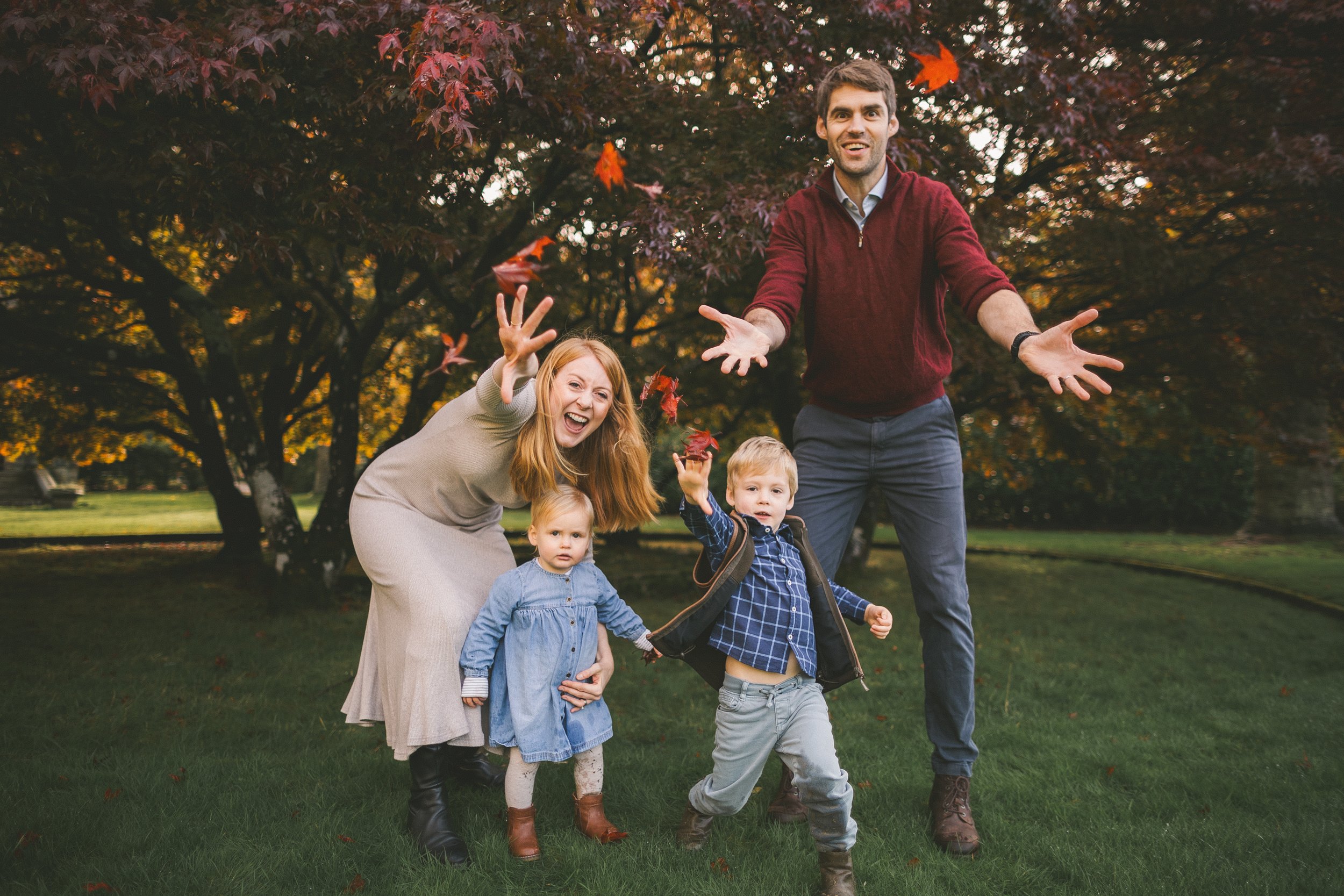 A family of five, including a father, mother, and three young children, enjoying a fall day outdoors with trees and falling leaves in the background.