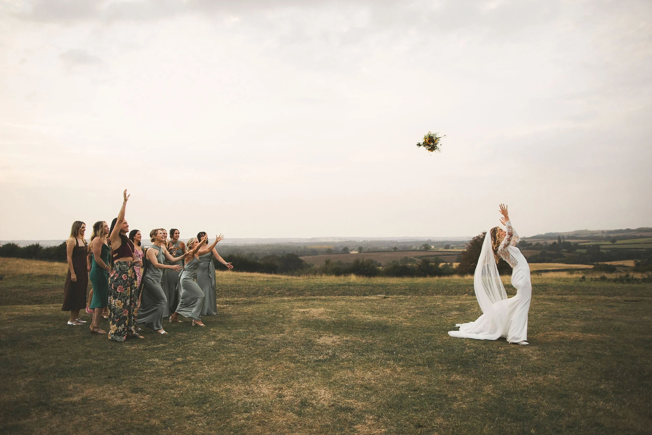 A bride in a white wedding dress throws a bouquet into the air at an outdoor wedding ceremony, with a group of women reaching out to catch it on a grassy field with a cloudy sky.