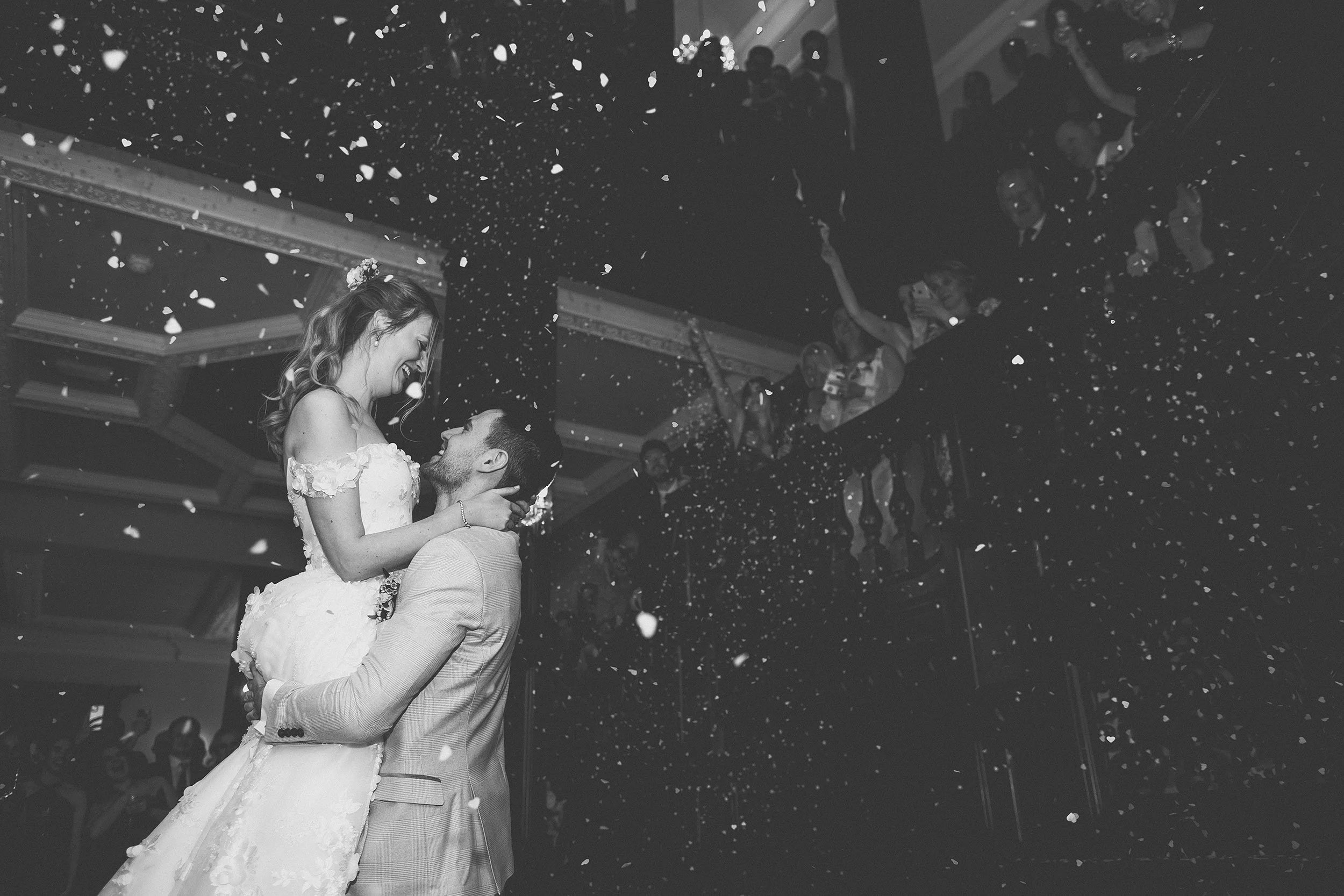A bride and groom dancing at their wedding reception, with confetti falling around them and guests in the background.