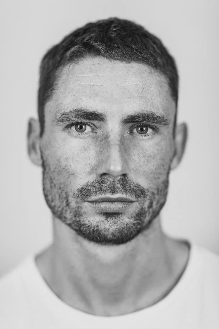 Black and white close-up portrait of a man with short hair and a beard, looking directly at the camera.