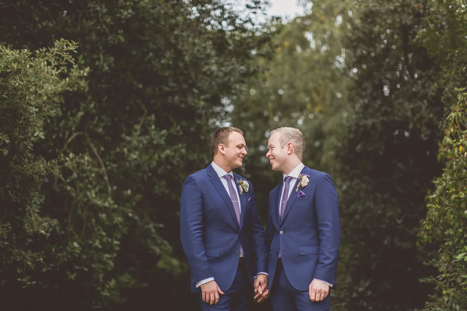 Two men in blue suits holding hands and smiling at each other outdoors with green trees in the background, celebrating their wedding.
