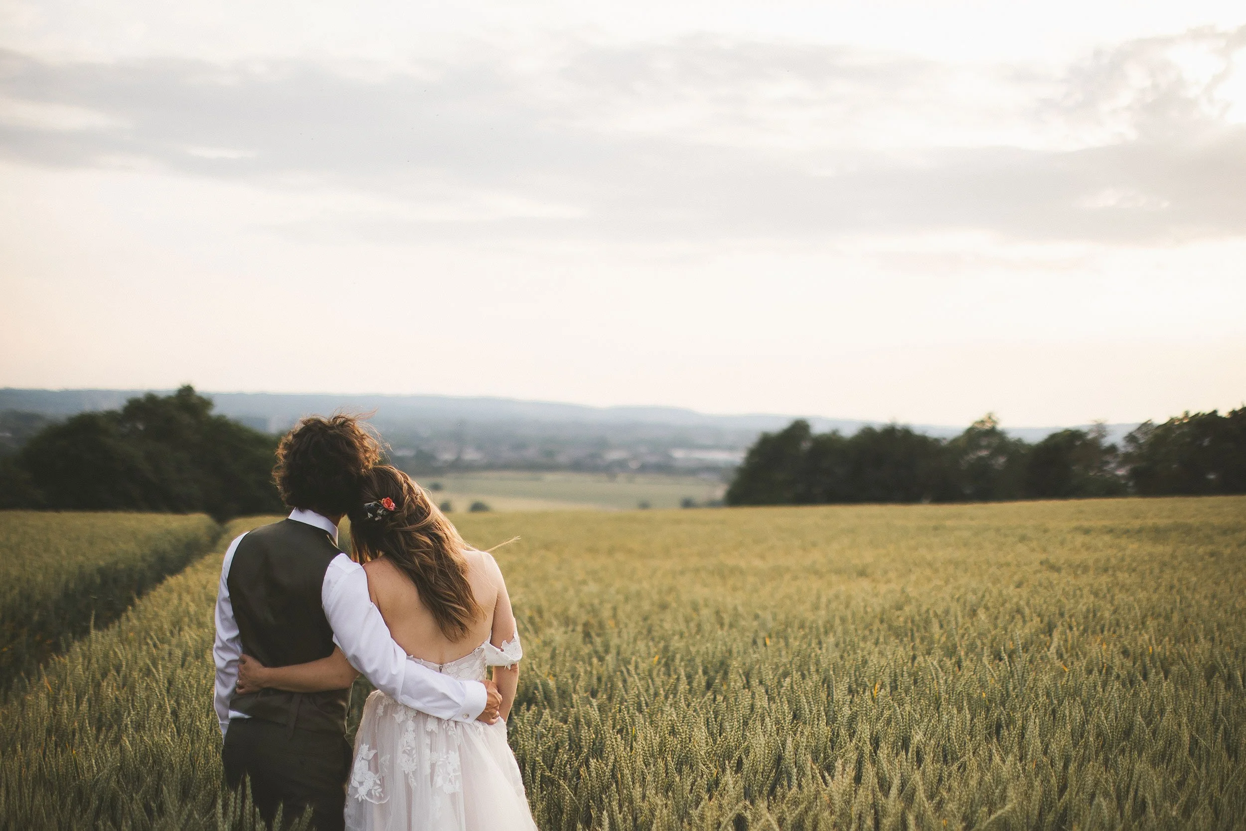 A couple dressed in wedding attire standing close together in a field, facing away and holding each other, with a scenic landscape and cloudy sky in the background.