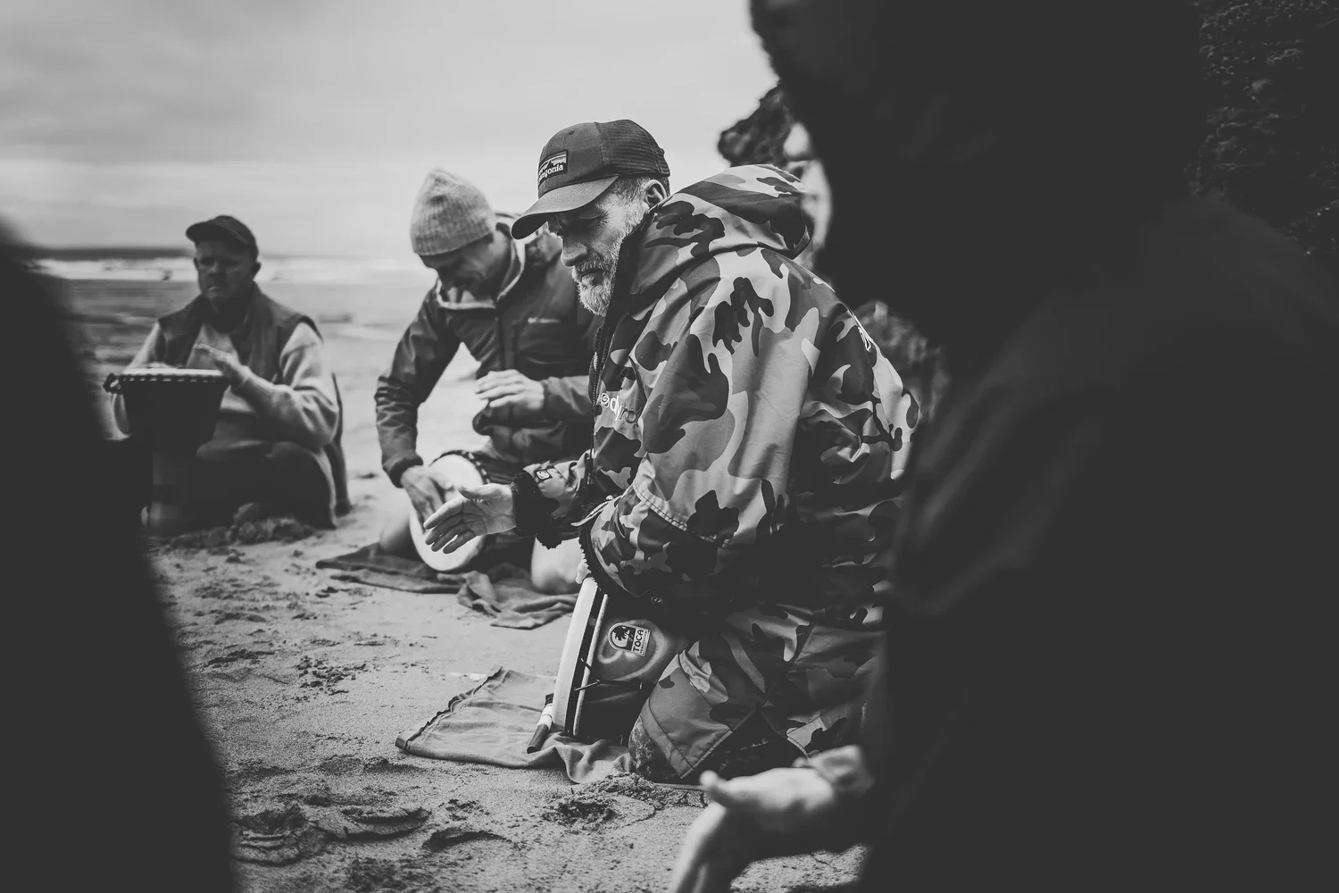 Group of people praying on a beach, some kneeling with prayer mats, black and white photo.