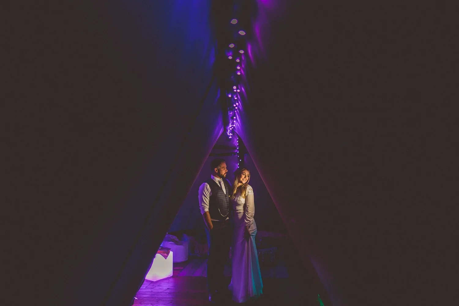 A couple is standing in a dark tunnel with purple and blue string lights overhead. The woman is smiling and wearing a white dress, while the man is looking at her and wearing a vest and shirt.