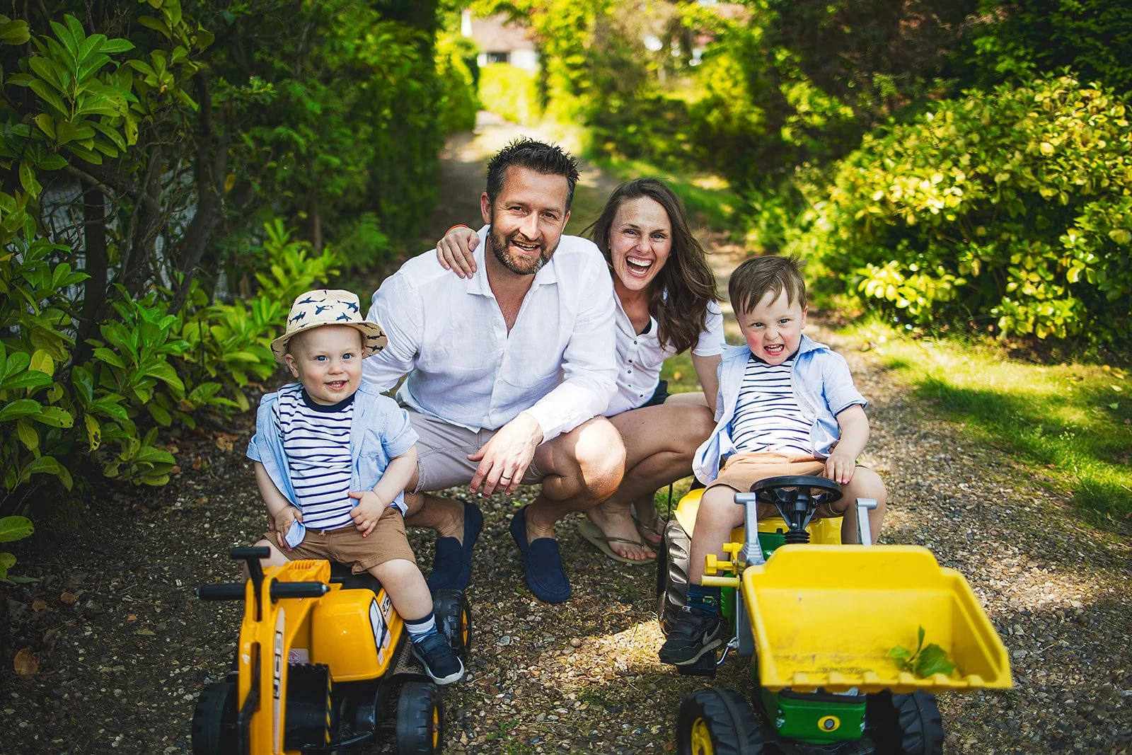 A happy family consisting of two children and two adults, posing outdoors on a sunny day among green foliage. The children are sitting on toy trucks, smiling, with the adults kneeling behind them, also smiling and enjoying the moment.