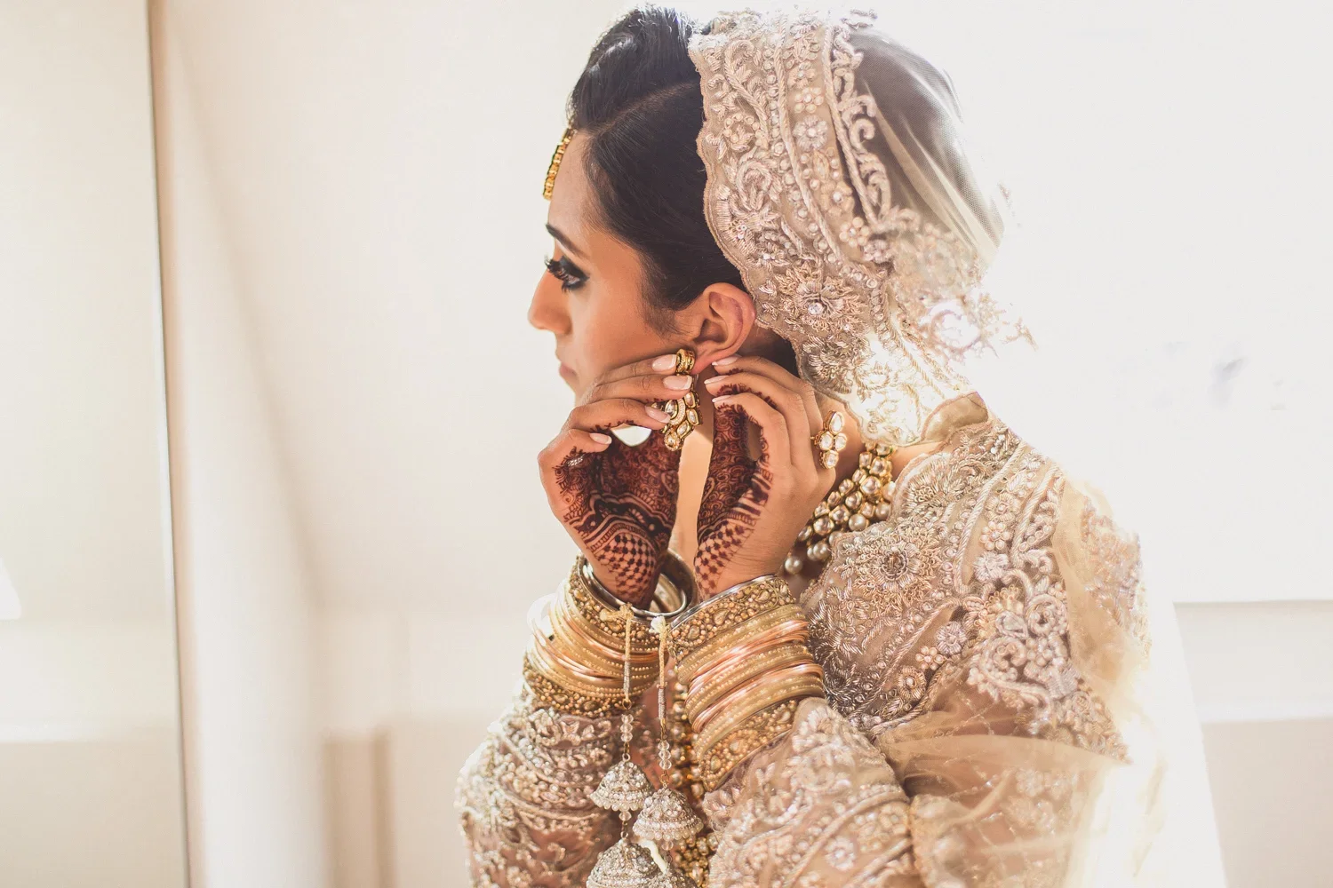 A woman in traditional Indian bridal attire adjusting her earring, adorned with jewelry, bangles, and mehndi on her hands, with a richly embroidered outfit and head covering.