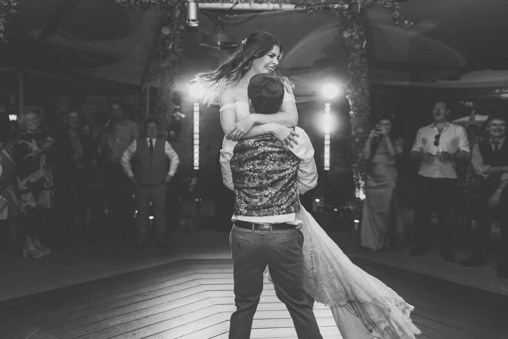 A black-and-white photo of a couple dancing closely during a wedding reception, with guests watching and smiling in the background.