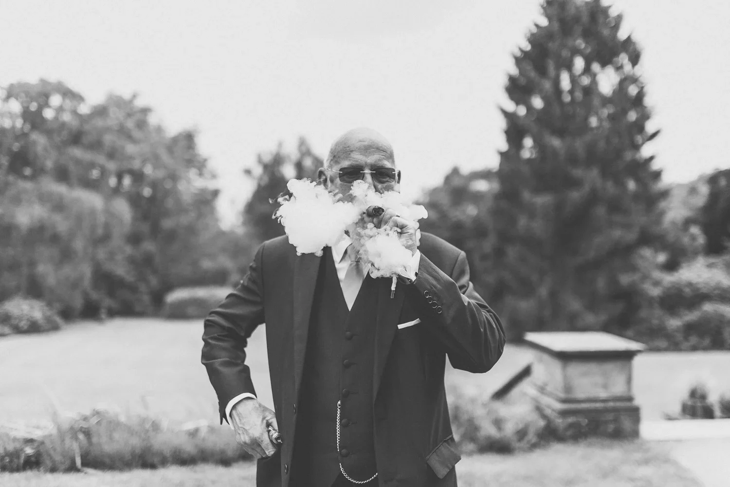 A man wearing a suit and sunglasses is smoking a cigar outdoors, with smoke around his face and a garden with trees in the background.