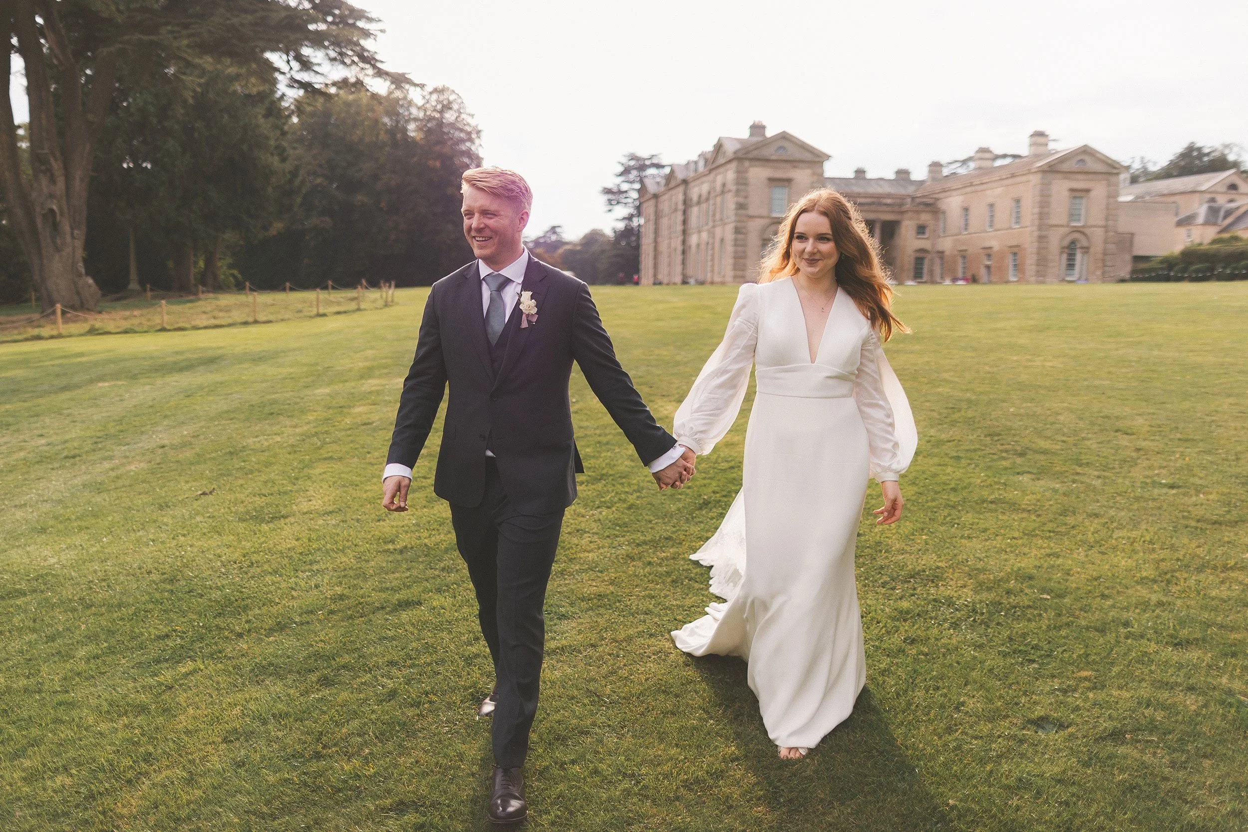 A happy couple in wedding attire walking hand in hand on a lush green lawn in front of a grand historic building.