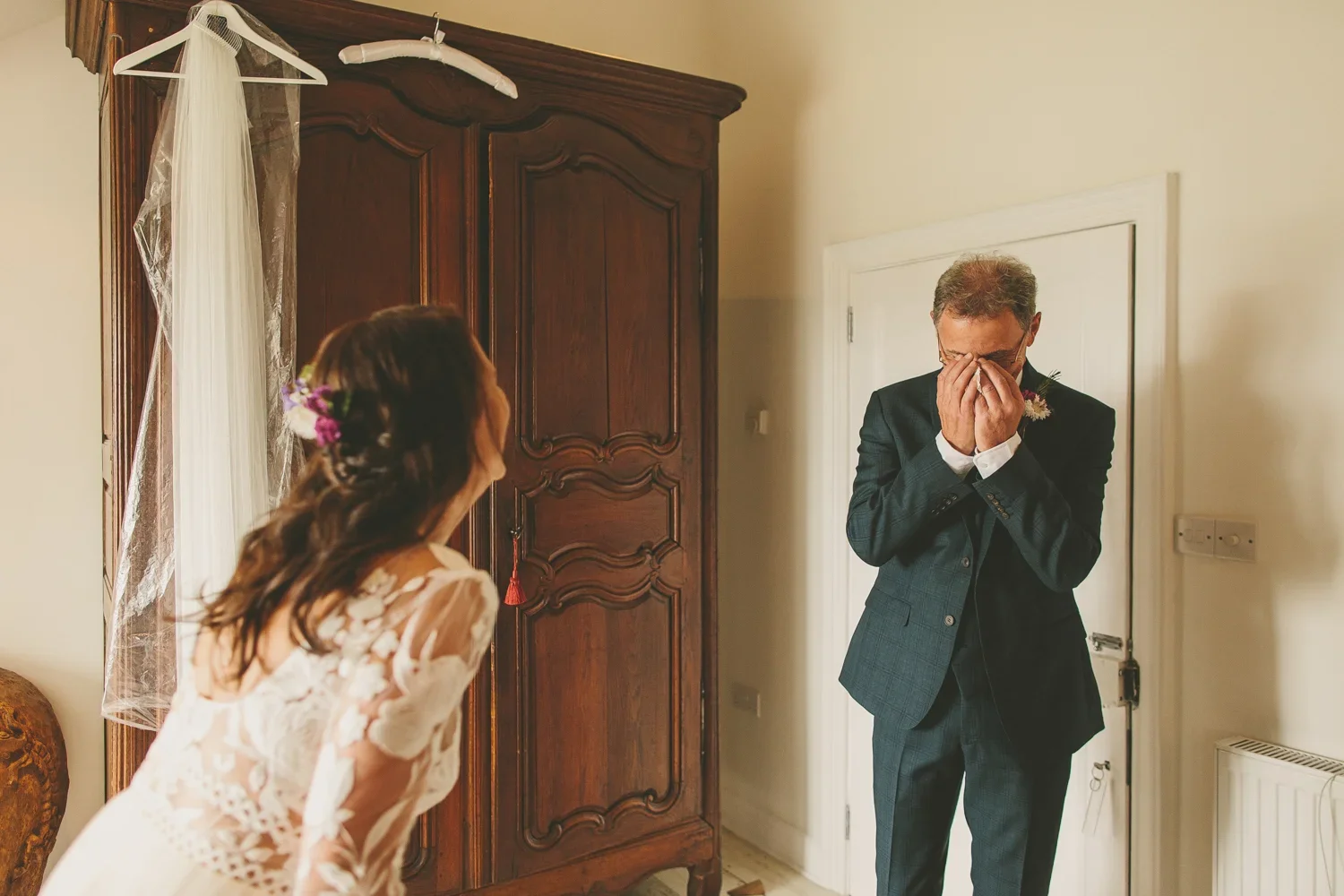 A bride with brown hair and purple flowers in her hair leans forward in front of an emotional groom who is wiping his eyes, in a room with a wooden armoire and a white door.