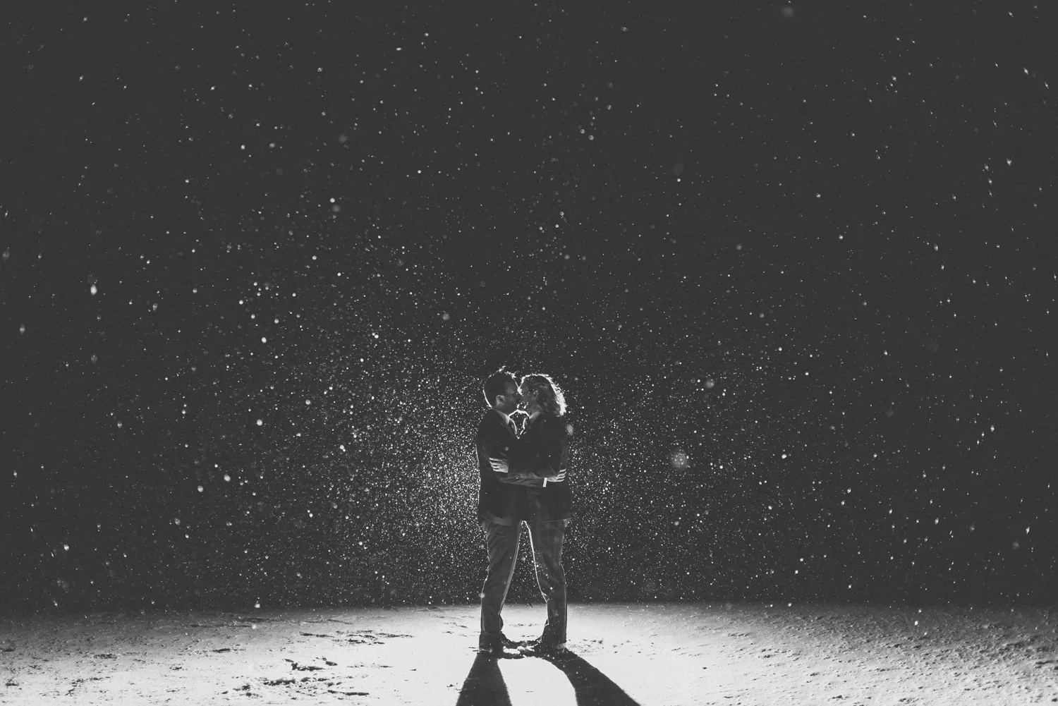 A black and white photo of a couple standing close together on a beach at night, with rain falling around them and their shadows cast on the sand.