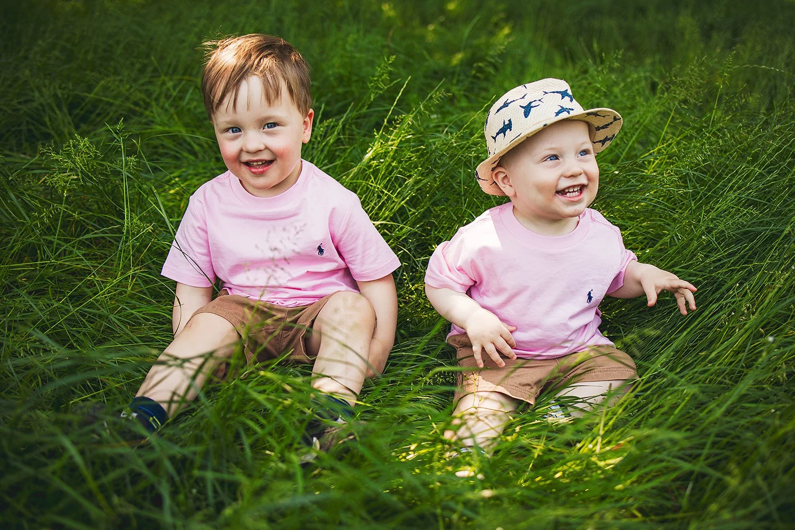 Two young boys sitting in tall grass outdoors, smiling and enjoying nature, wearing pink shirts and khaki shorts; one boy has a hat with a shark pattern.