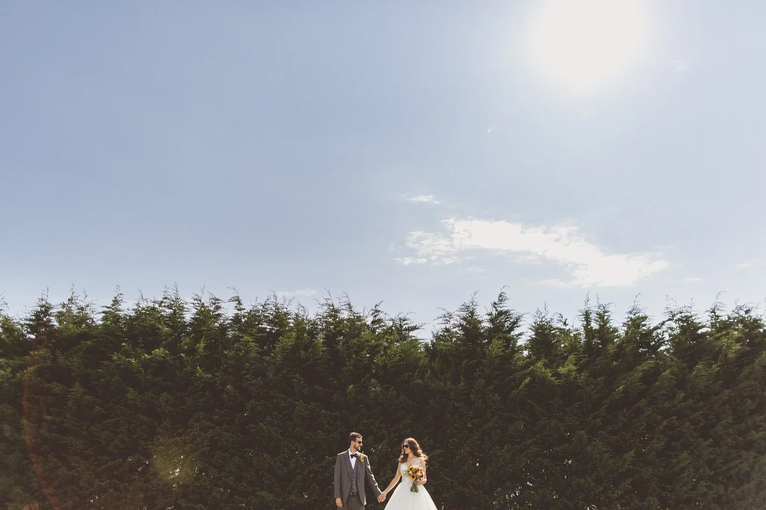 A bride and groom holding hands outdoors under a clear blue sky with a row of green trees behind them.