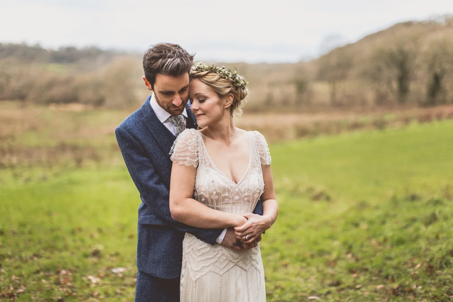 A newlywed couple embraces outdoors in a green field with trees in the background, the bride wearing an elegant lace wedding dress and the groom in a dark suit.