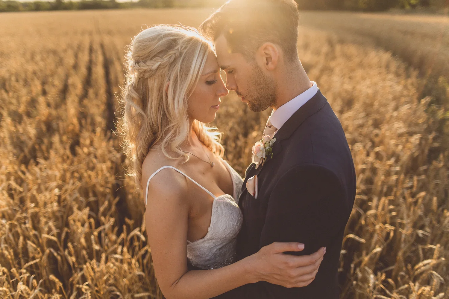A newlywed couple dressed in wedding attire in a field of wheat at sunset, with their foreheads touching and eyes closed.
