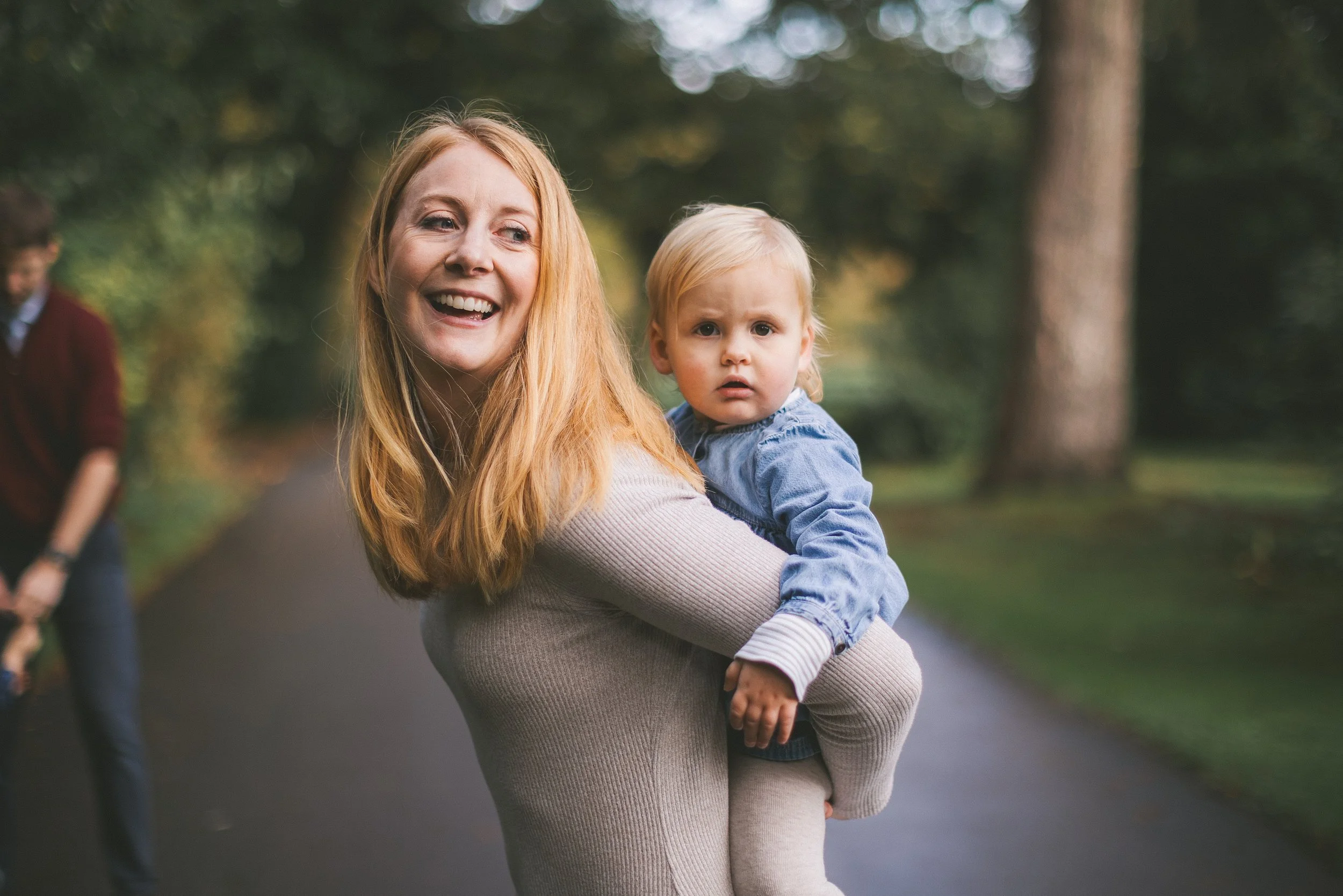 A red-haired woman smiling and holding a young blonde boy in her arms outdoors on a park path with trees in the background.