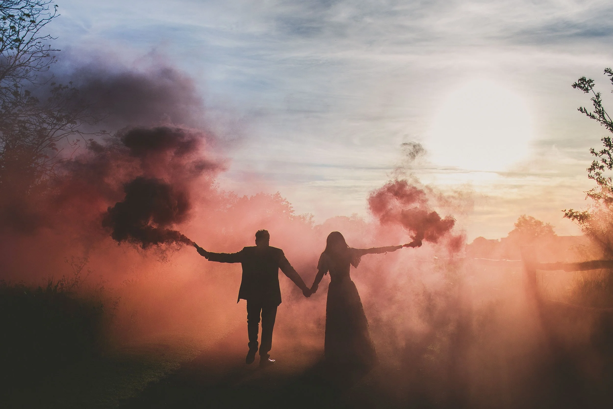 Two people are holding smoke bombs emitting pink and purple smoke, standing outdoors at sunset with trees on either side, creating a colorful hazy scene.