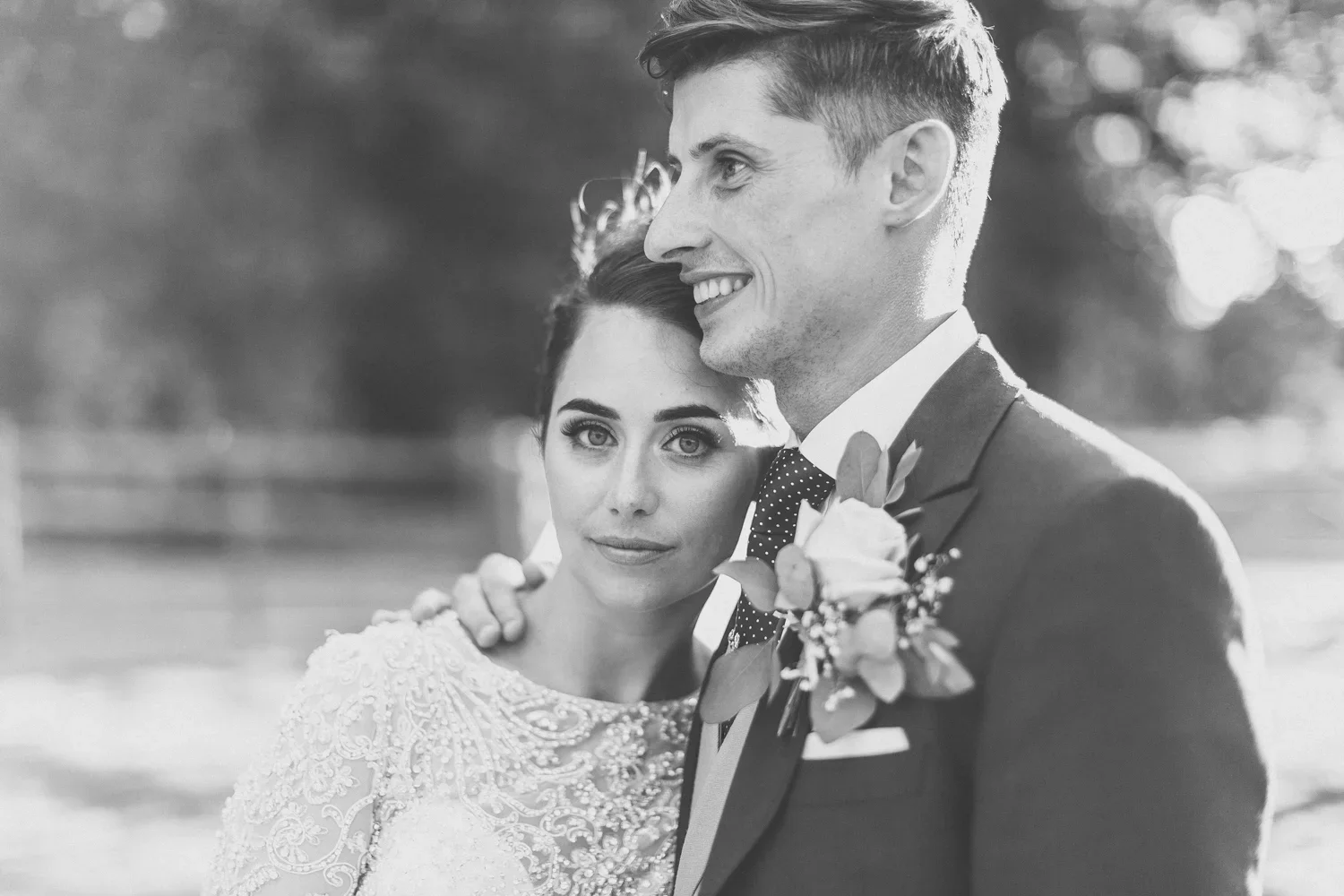 Black and white photo of a newlywed couple outdoors, with the bride looking directly at the camera and the groom smiling, wearing a suit with a boutonniere.
