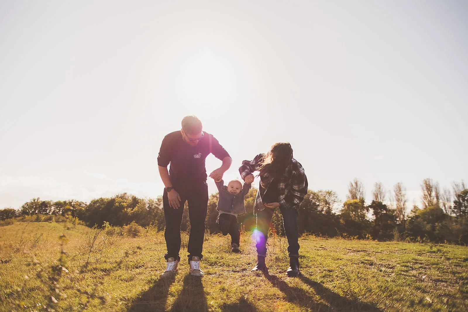 A family of three, a father, mother, and young child, enjoying outdoor time in a grassy field during sunset, with the child being lifted between the parents.