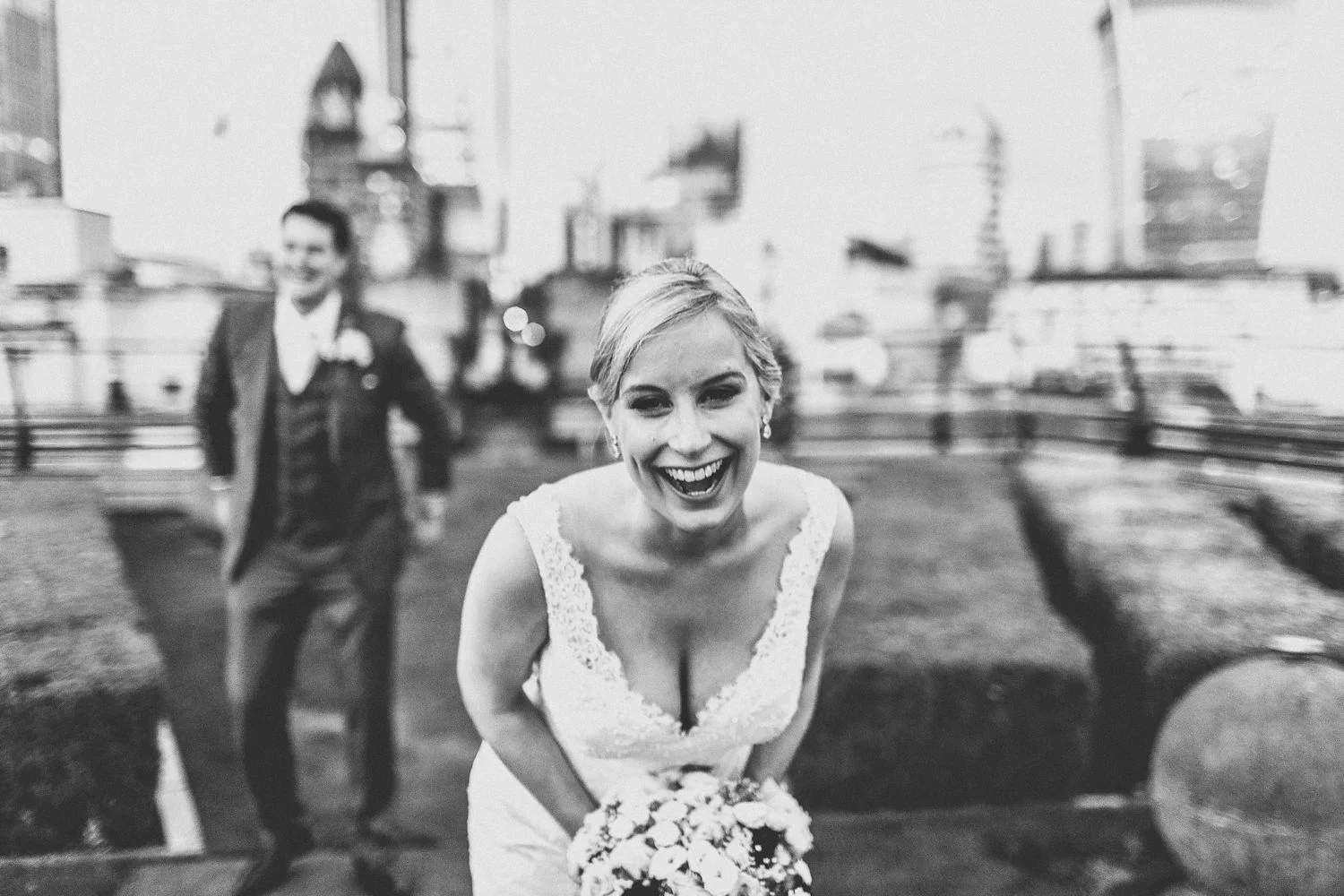 Black-and-white photo of a smiling bride holding a bouquet, with a groom in a suit blurred in the background, on a rooftop with city buildings.