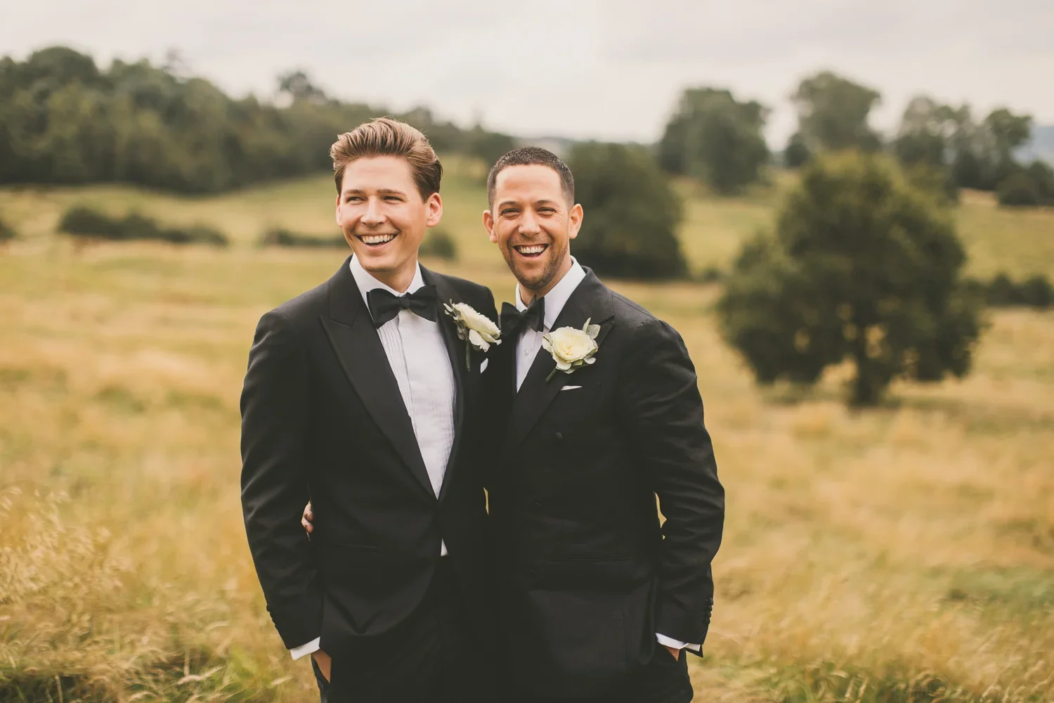 Two men dressed in black tuxedos with bow ties and white shirts, standing outdoors in a grassy field with trees in the background, smiling and embracing each other, one with his hand in his pocket.