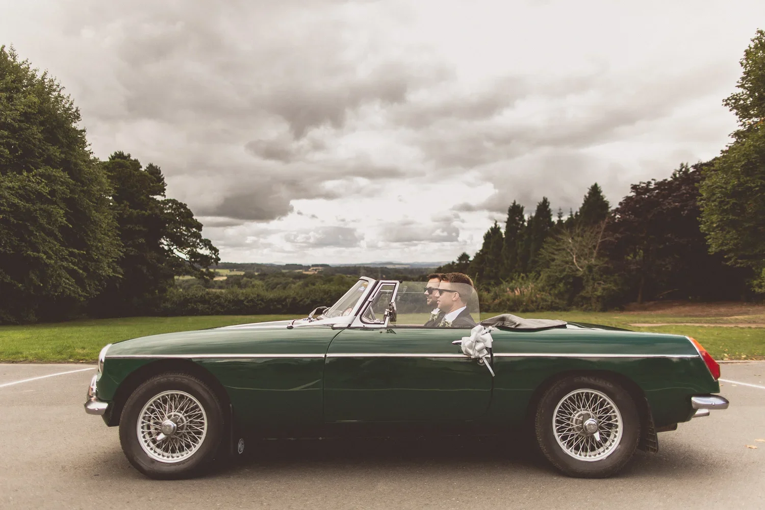 A vintage dark green convertible car parked outdoors on a paved surface with two men sitting inside, dressed in formal attire, possibly a wedding or special occasion, with trees and cloudy sky in the background.