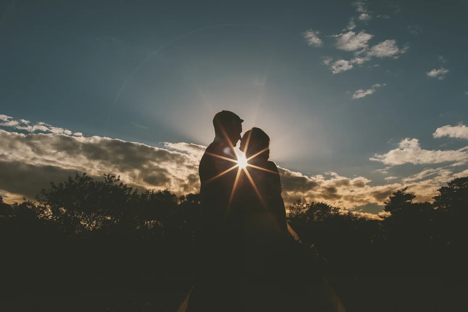 Silhouette of a couple facing each other at sunset with the sun behind them creating a starburst effect, trees and clouds in the background.