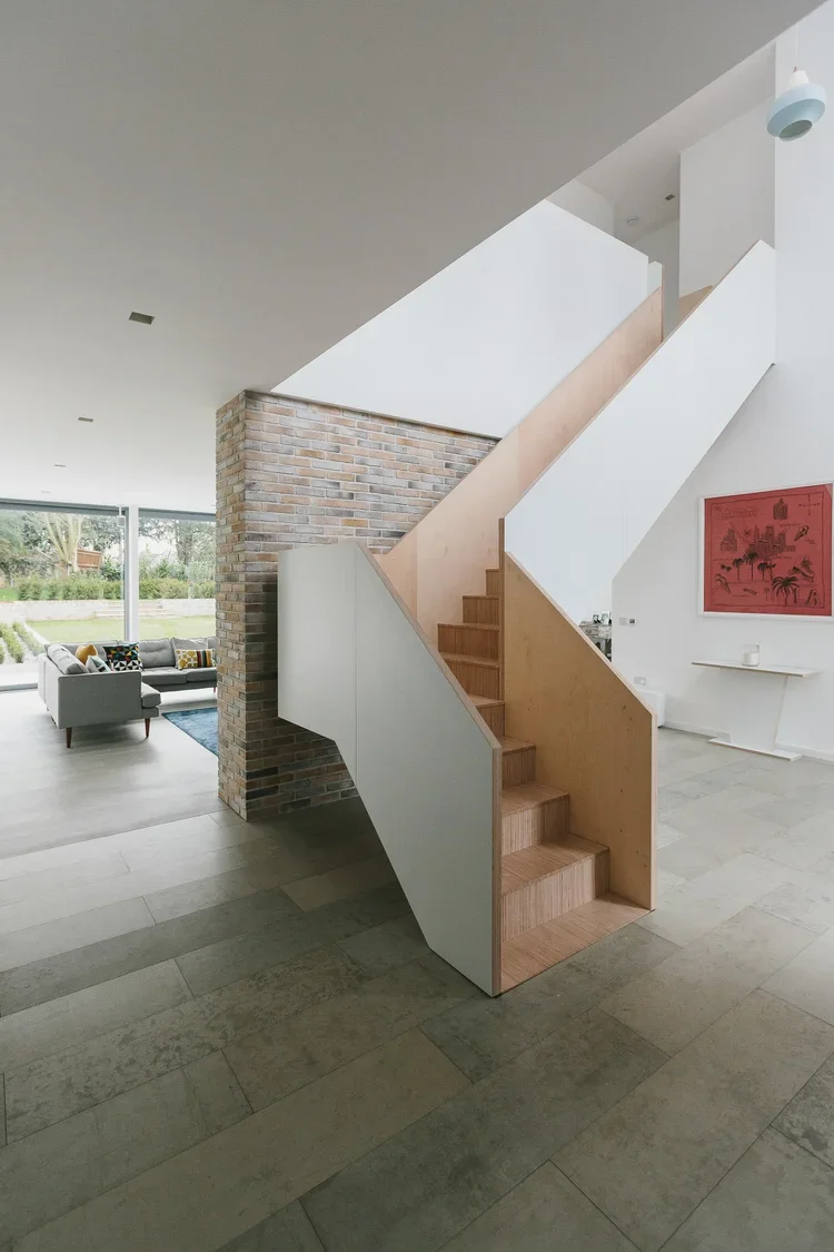 Modern interior with a staircase featuring wooden steps and a minimalist white and wood design, behind a brick wall, in a bright living space with large window and seating area.