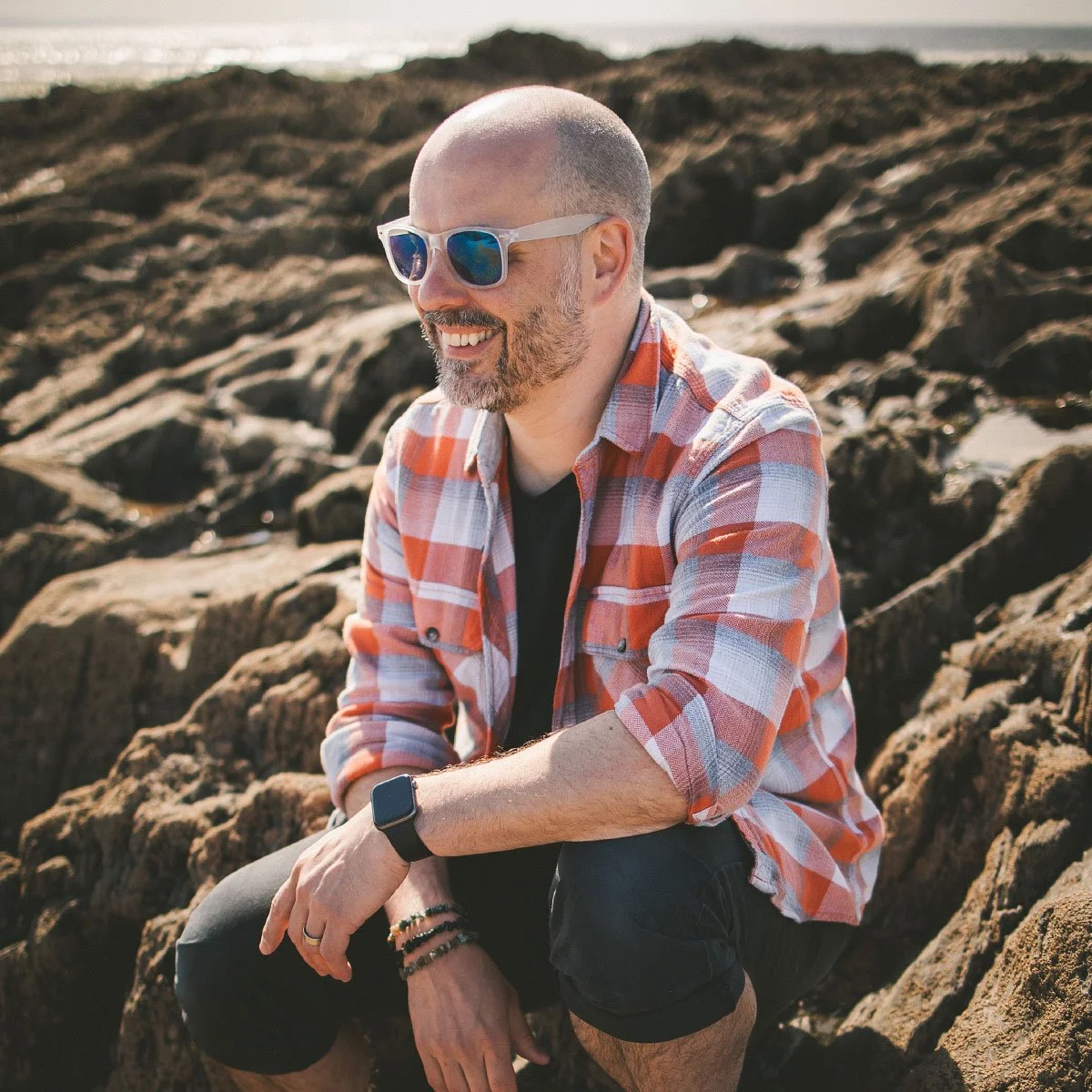 A man with a bald head, beard, and sunglasses sitting on rocks at the beach, smiling and looking to the side.