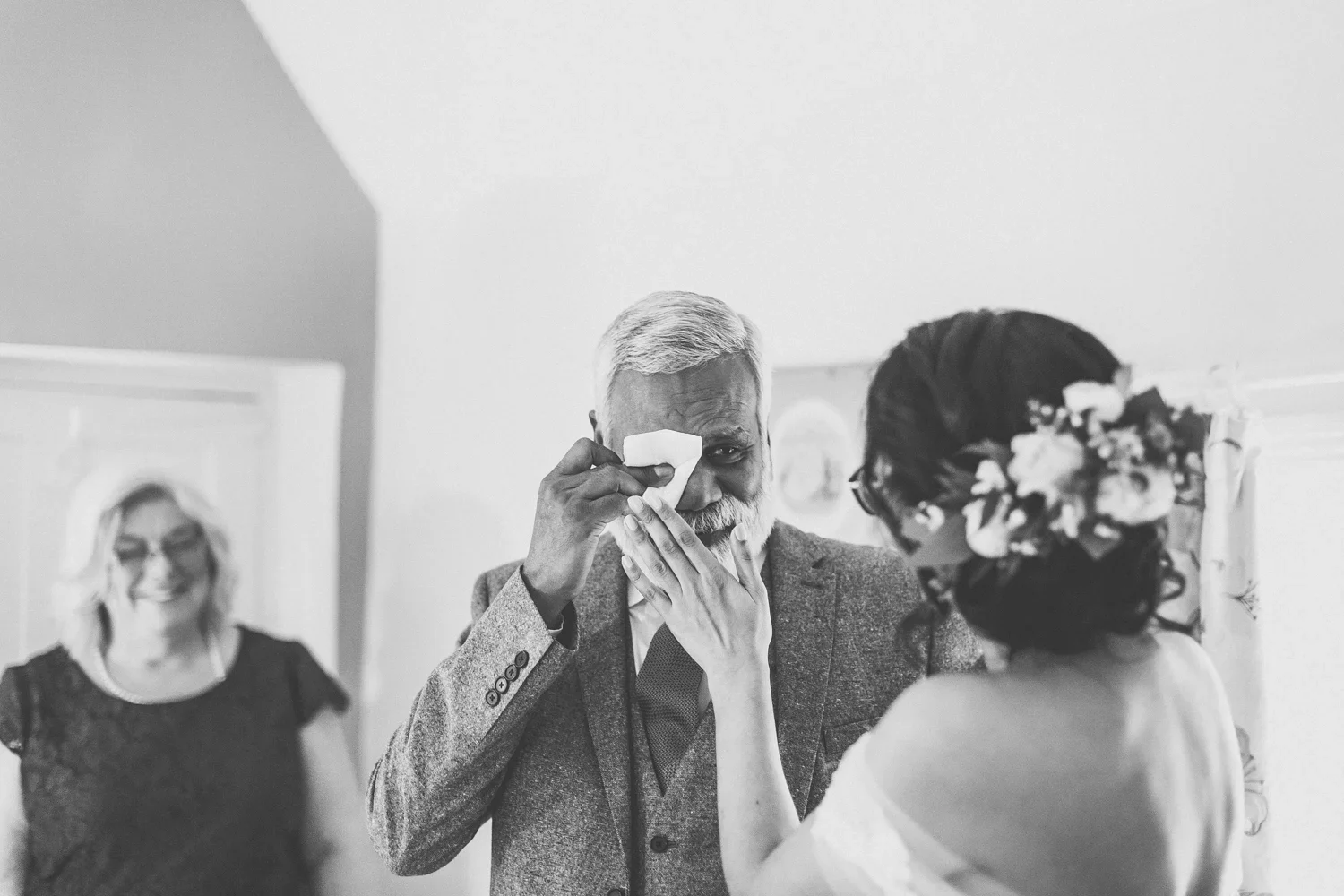 An elderly man with gray hair and a beard wiping tears with a tissue while a woman with flowers in her hair gently touches his face. A smiling woman in the background.