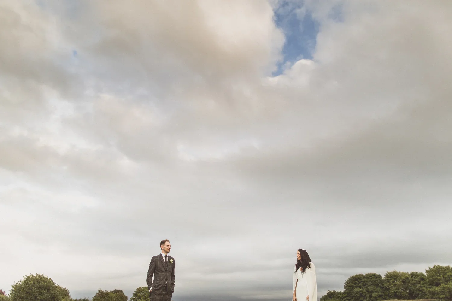 A man in a gray suit and a woman in a white dress standing apart outdoors under a cloudy sky, with trees in the background.