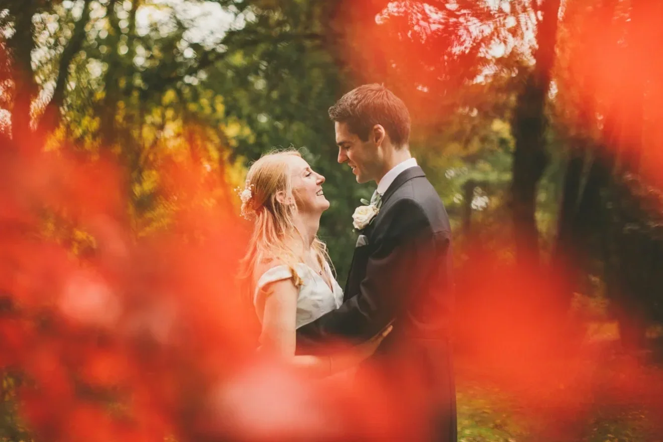 A bride and groom smiling at each other, standing outdoors in a wooded area with autumn leaves, framed by red-orange leaves in the foreground.
