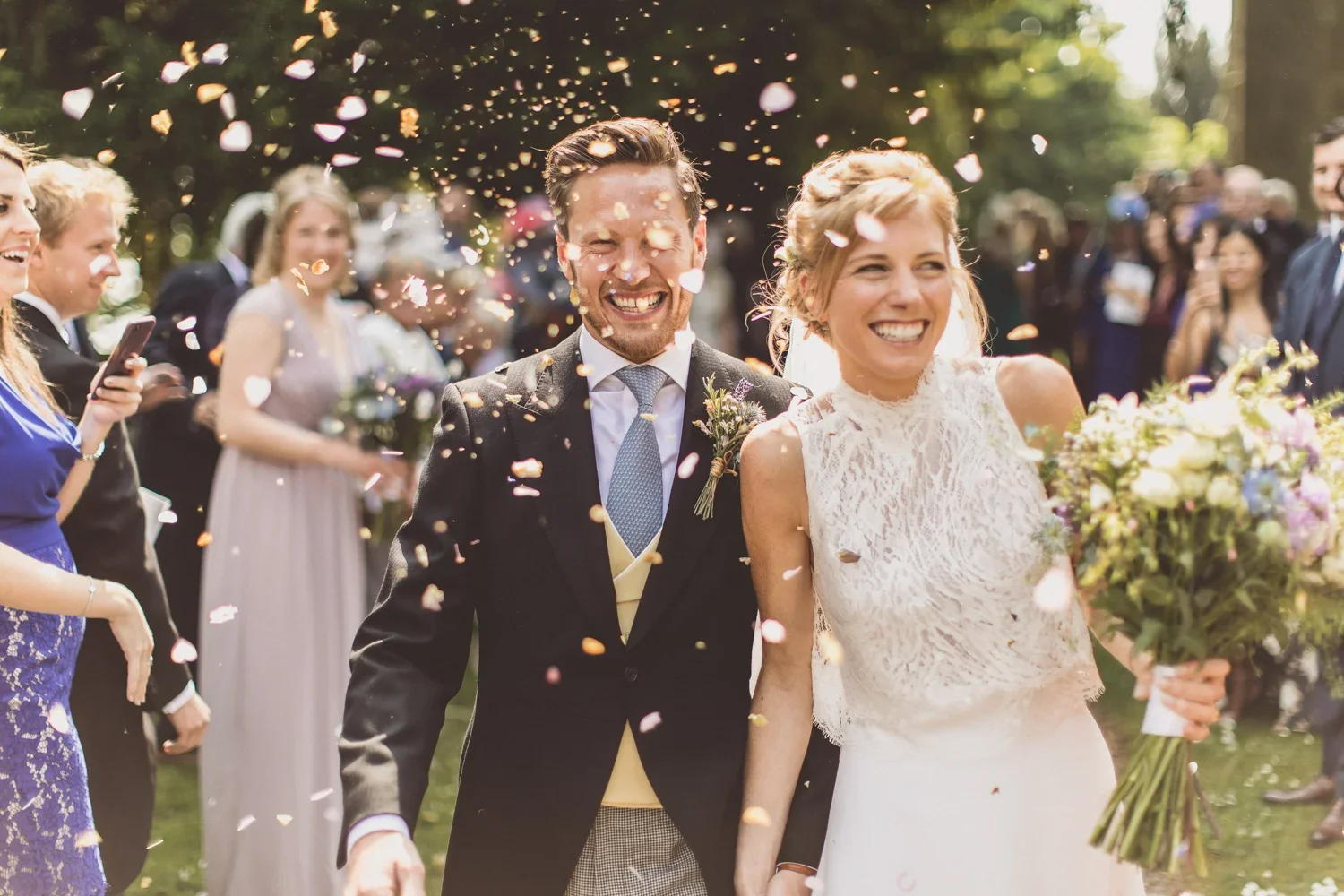 A bride and groom smiling as they walk together at their wedding, surrounded by friends and family celebrating with confetti and flowers outdoors.
