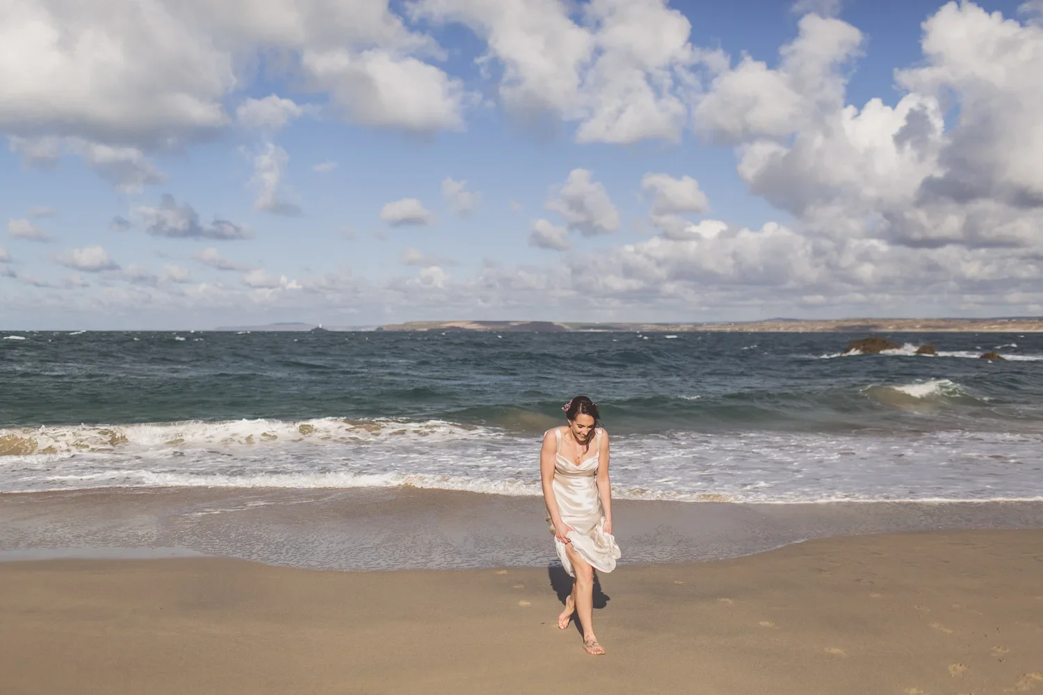 A woman in a beige dress is walking barefoot on the sandy beach near the shoreline, with ocean waves and a partly cloudy sky in the background.