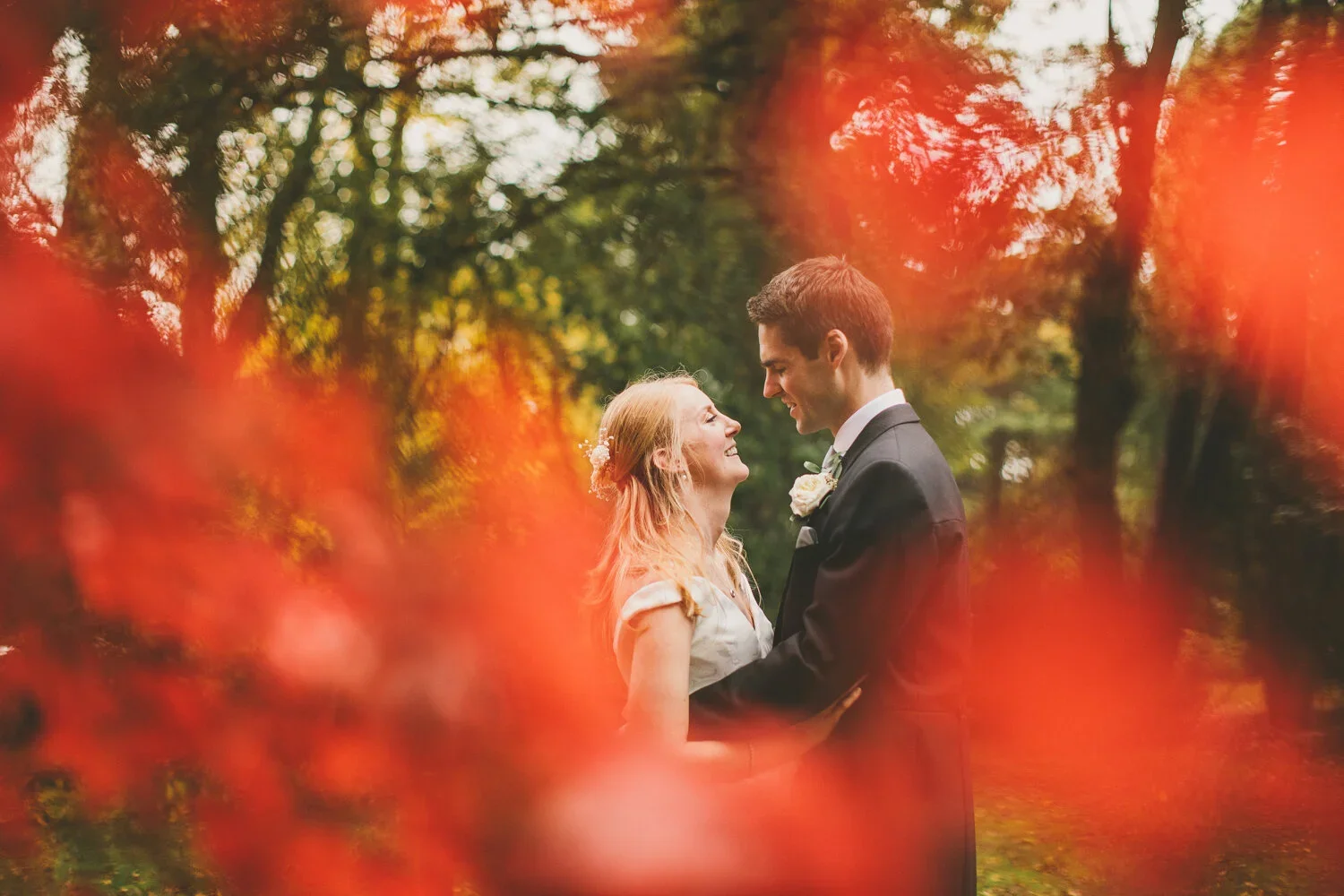 A couple in wedding attire standing close together outdoors surrounded by autumn trees with orange and yellow leaves, smiling and gazing at each other.