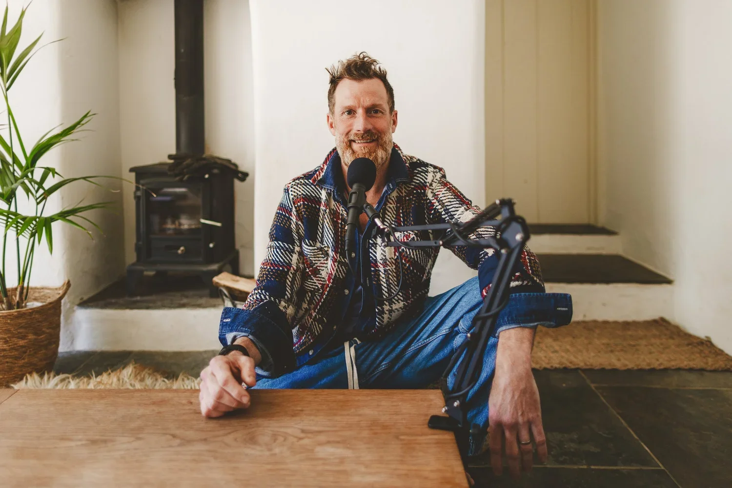 A man sitting cross-legged at a wooden table, smiling, with a microphone in front of him, in a cozy room with a wood stove, a plant, and simple decor.