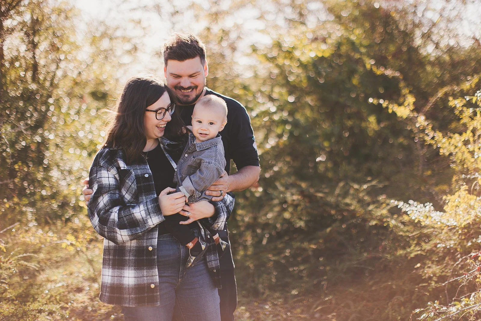 A smiling family of three, including a woman, man, and toddler, enjoying a walk outdoors in a park with trees and sunlight filtering through the leaves.