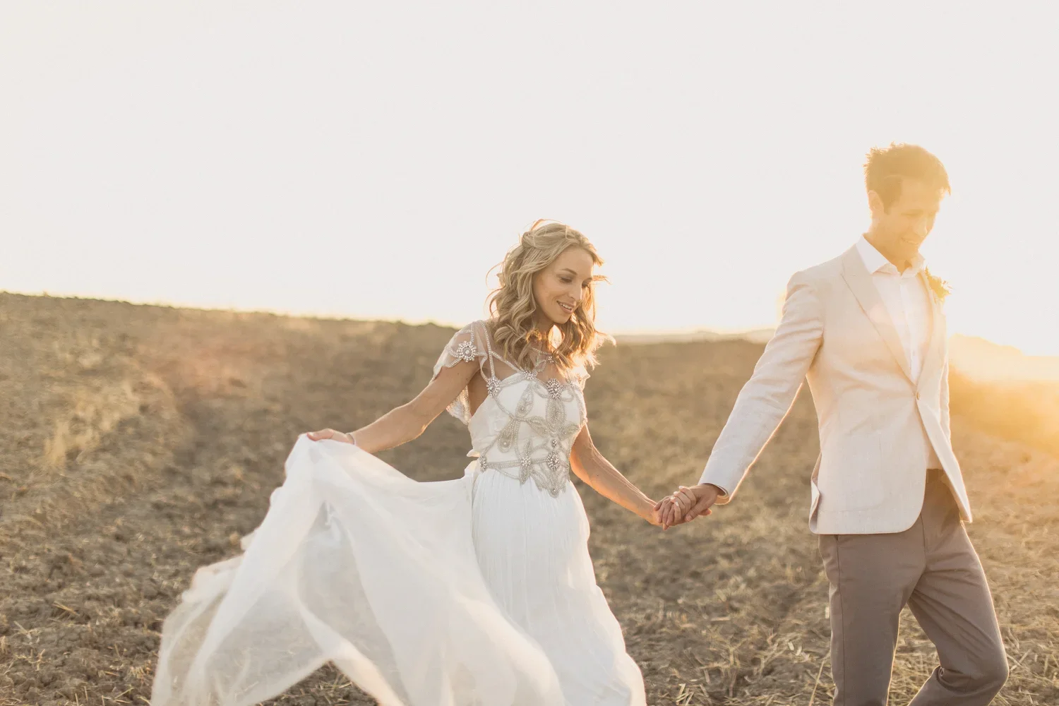 A bride and groom walking hand in hand through a field at sunset, with the bride lifting her dress and smiling.