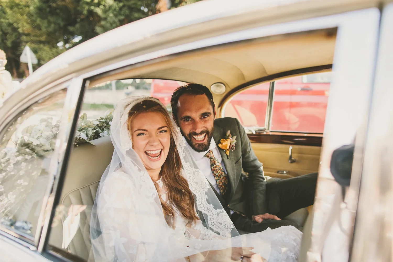 A newlywed couple sitting in the back of a vintage car, smiling and laughing, with greenery and a red vehicle outside.