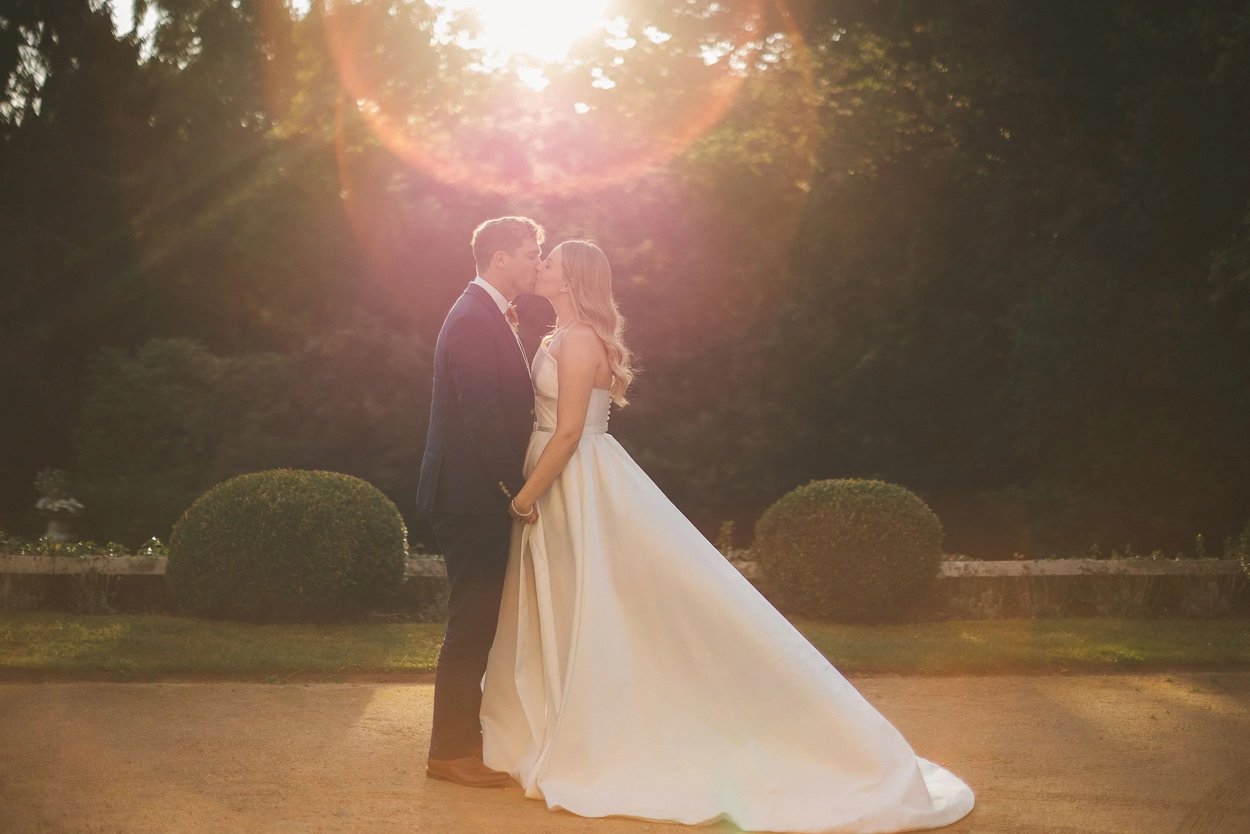 A bride and groom sharing a kiss outdoors during sunset, with trees in the background and the bride wearing a white wedding dress and the groom in a dark suit.