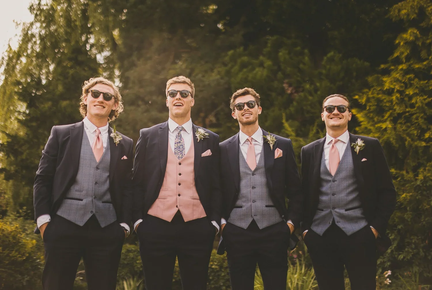 Four men dressed in suits and ties, wearing sunglasses, smiling outdoors with trees in the background.
