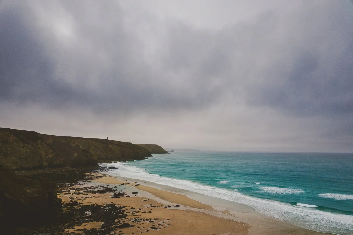 Coastal scene with sandy beach, rocky cliffs, and ocean under a cloudy sky.