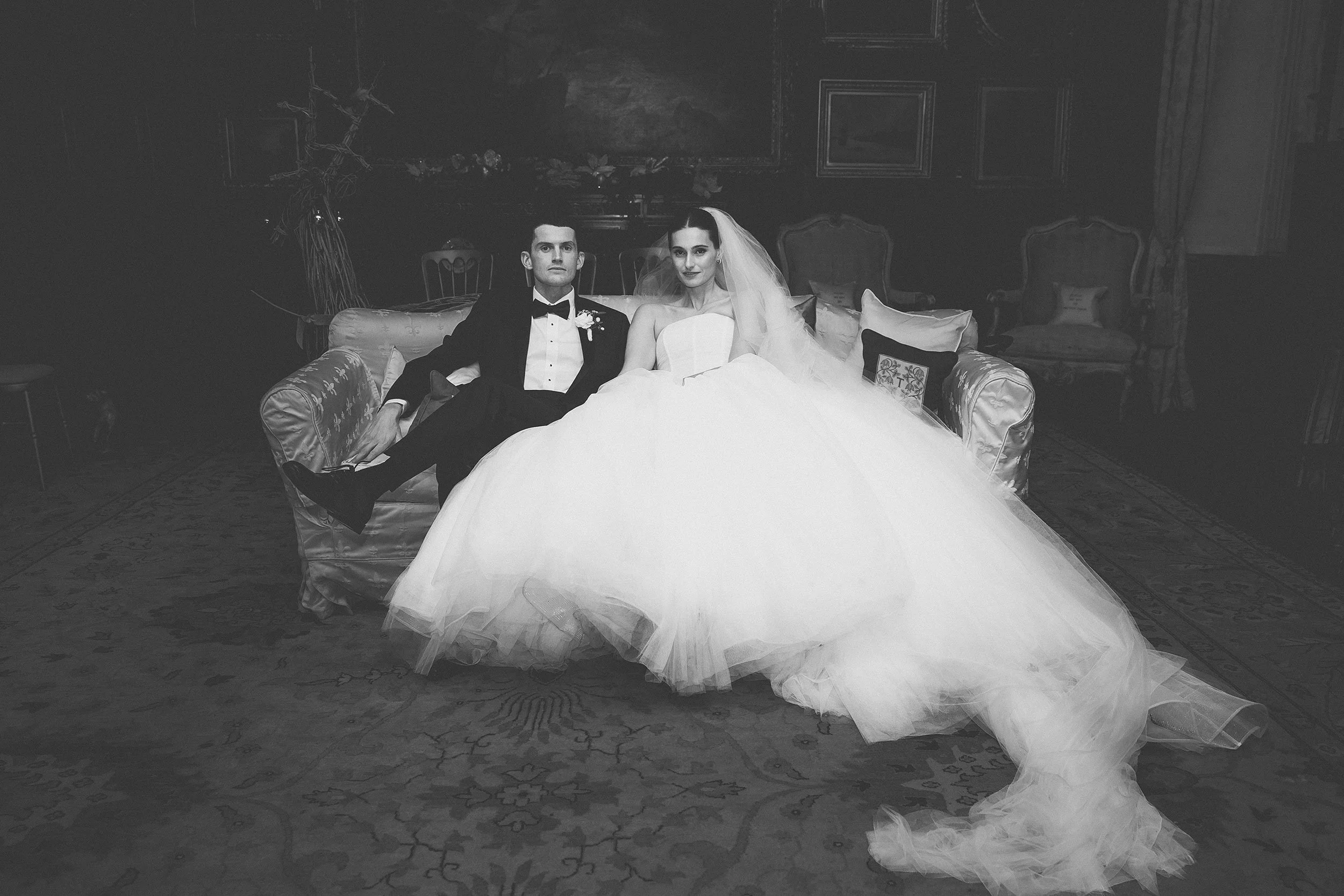 Black and white photo of a bride and groom sitting together on a sofa in a decorated room. The groom is dressed in a tuxedo, and the bride is wearing a wedding gown with a veil. The room has ornate chairs and artwork on the walls.