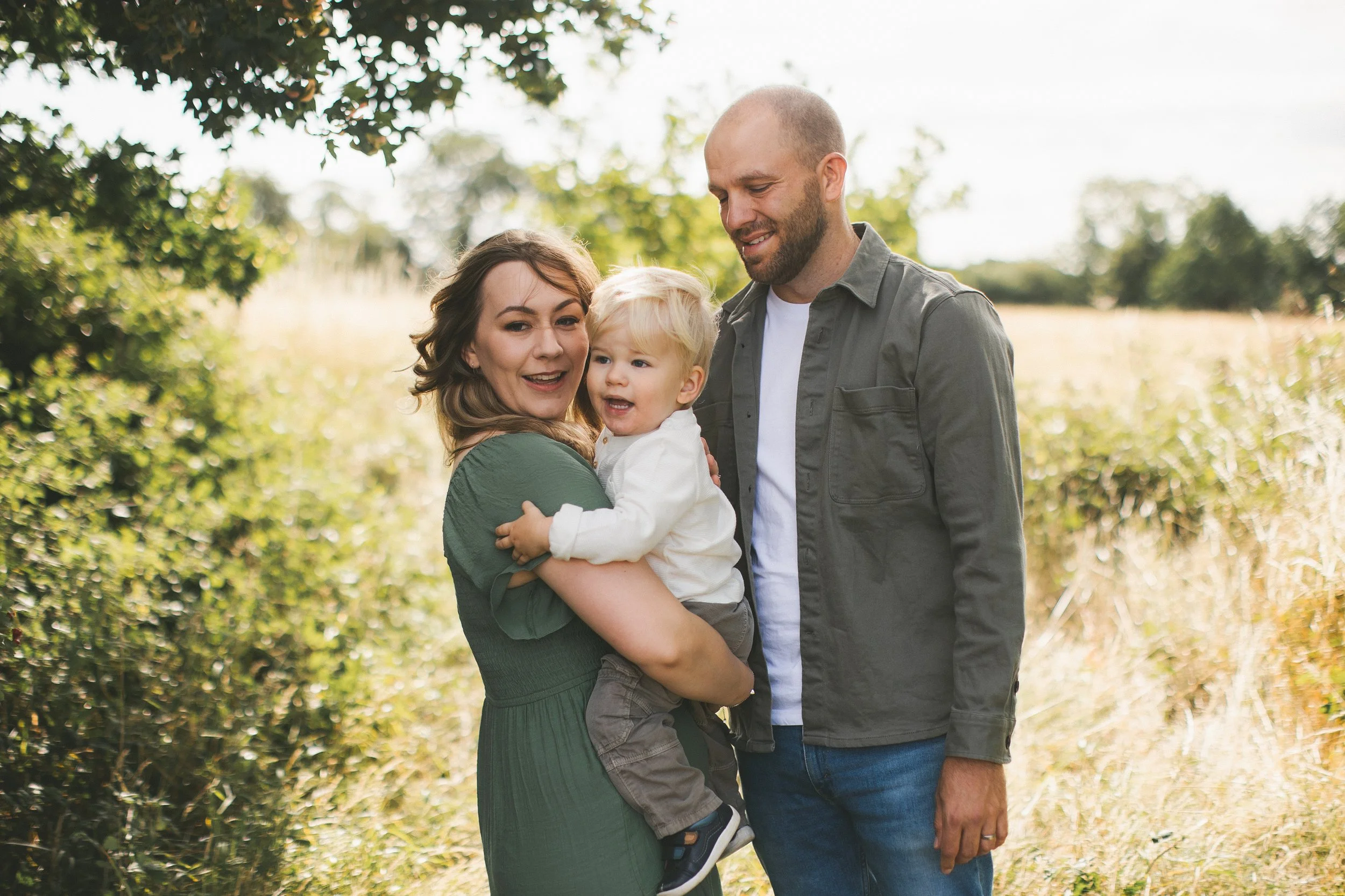A smiling woman holding a young boy while a man stands beside them outdoors in a sunny, green setting.