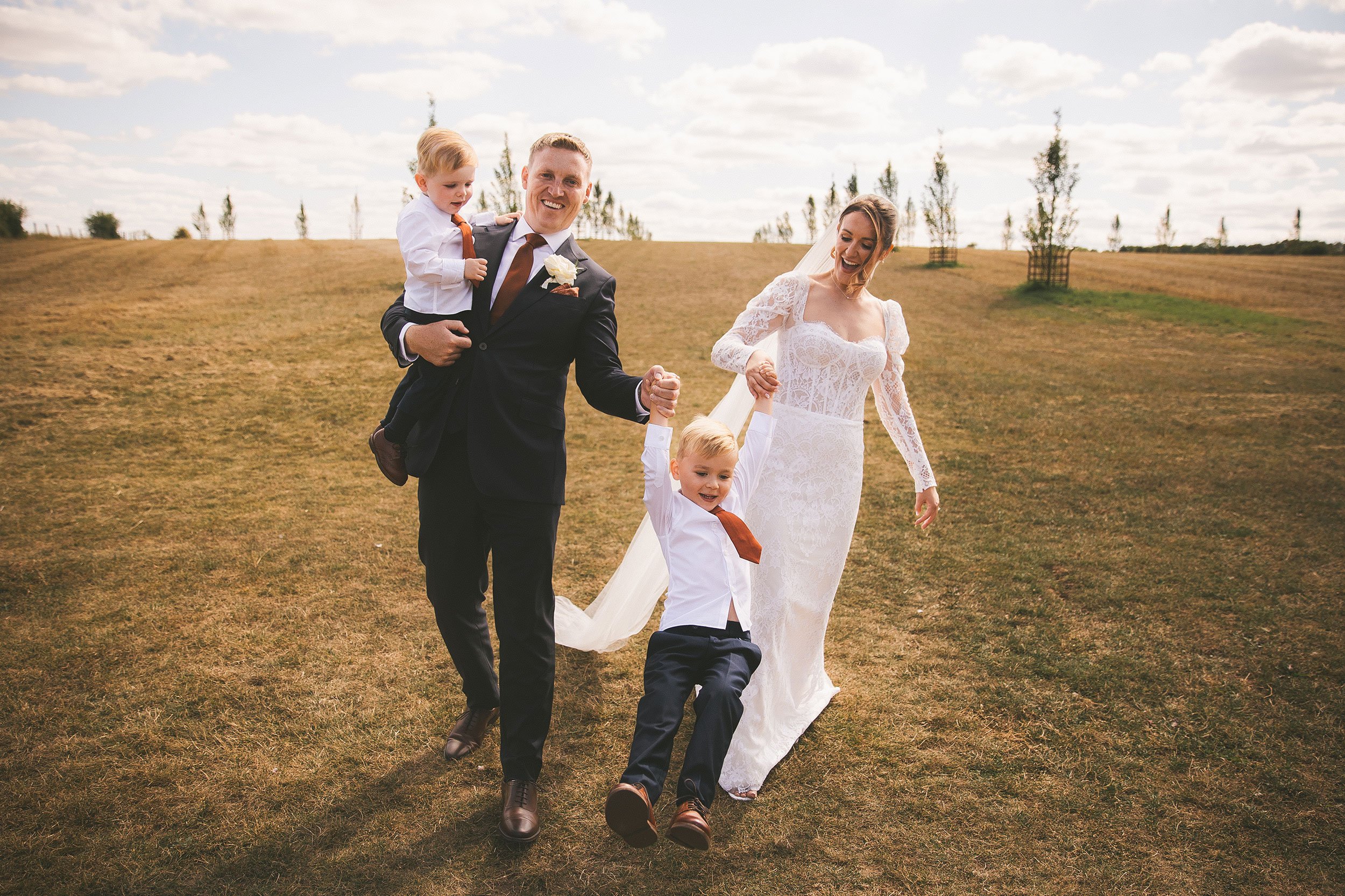 A bride and groom walking outdoors on a grassy field, holding hands with two young boys. The groom is carrying a boy on his shoulder, and all are smiling. The bride is wearing a white lace wedding dress, and the groom is in a dark suit with a white shirt and tie. The scene is set on a partly cloudy day with small trees in the background.
