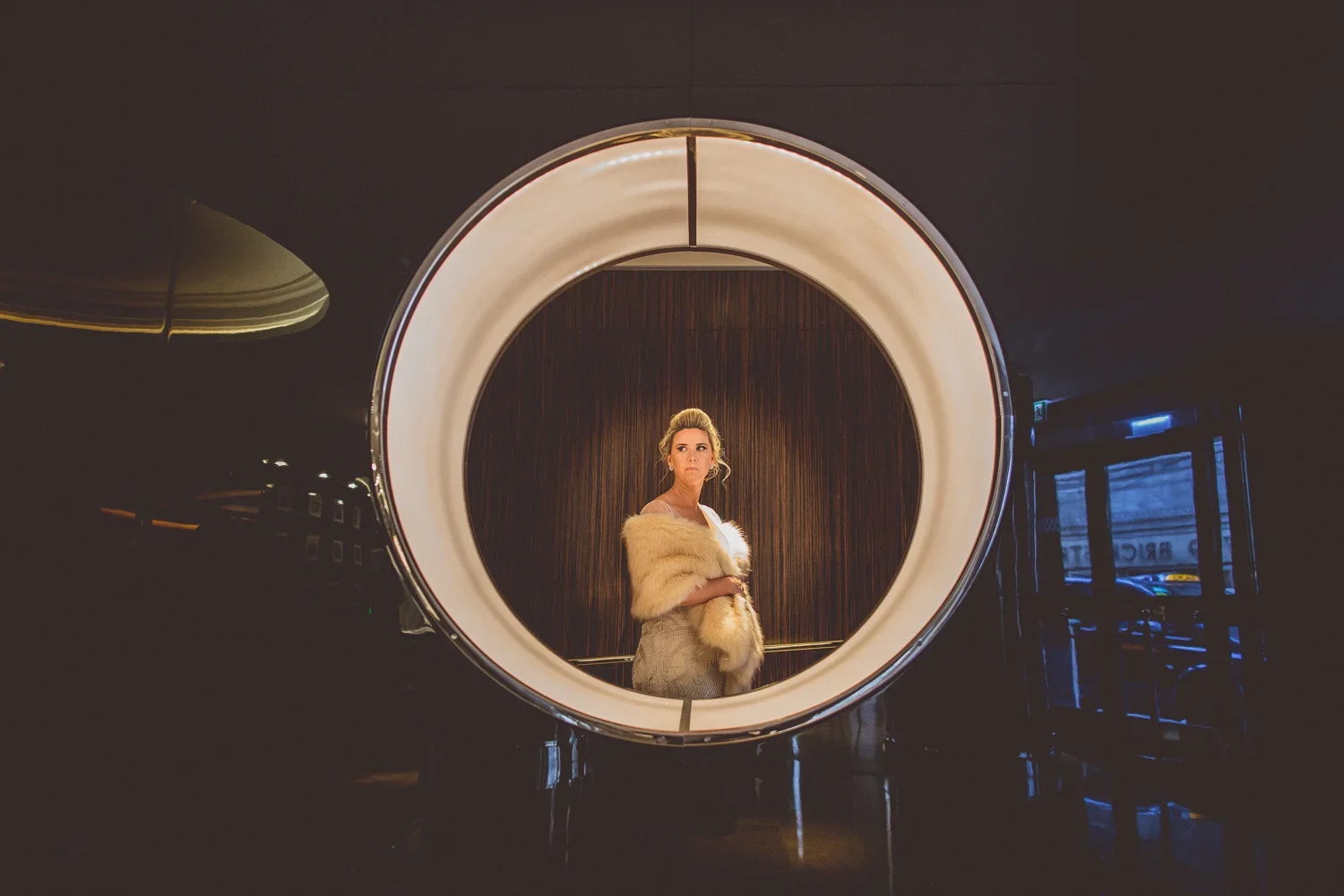 A woman standing inside an illuminated circular frame, dressed in a formal gown and fur stole, with a wood-paneled background.
